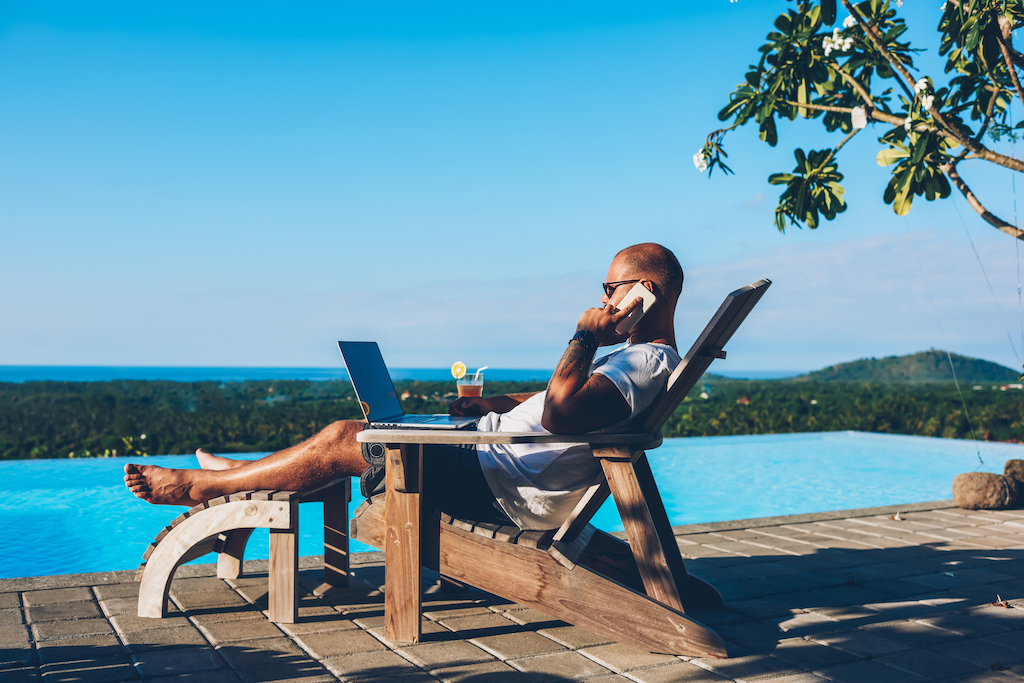 Young businessman resting on sunbed enjoying summertime in resort spa near swimming pool while working remotely at laptop computer connected to wireless internet and talking on smartphone with partner